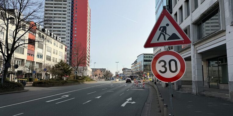 Blick auf die Kreuzung. Rechts: ein rotes Dreieck-Warnschild mit dem Symbol eines Arbeiters (Baustelle) und ein rundes weißes Schild mit rotem Rand und der Zahl 30 (Geschwindigkeitsbegrenzung 30 km/h).